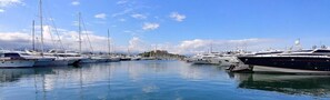 A marina with multiple yachts docked in calm waters, a clear sky, and a distant hill with buildings. - Hello Côte d'Azur - Spacious apartments at a 5 min walk from the beach (Antibes)