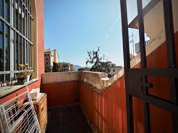 Un étroit couloir extérieur avec un mur rouge, un portail en métal et une fenêtre ornée de plantes en pot.
