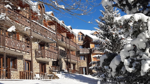 A multi-story wooden building with balconies, surrounded by snow-covered trees and a clear blue sky. - VVF Résidence Les Angles Pyrénées (Les Angles)
