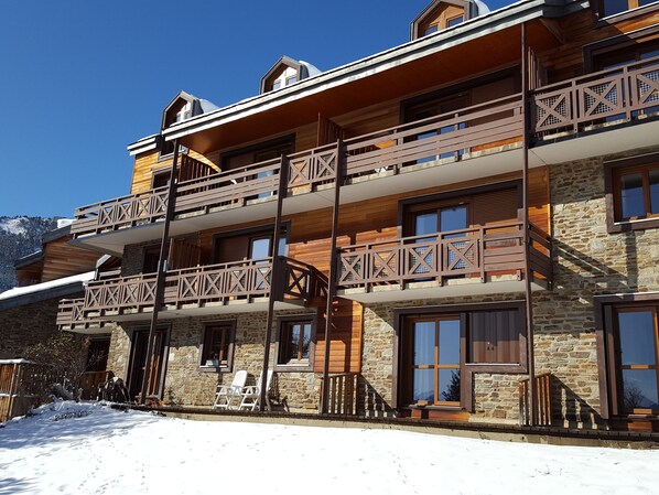 A multi-story building with wooden balconies and stone walls, featuring outdoor seating and a snowy landscape. - VVF Résidence Les Angles Pyrénées (Les Angles)