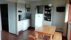 A compact kitchen with white cabinets, a microwave, a sink, and a refrigerator. There is a wooden table with benches and a television mounted on the wall. - VVF Résidence Les Angles Pyrénées (Les Angles)