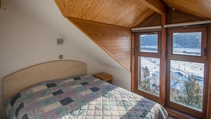 A bedroom with a wooden sloped ceiling, a bed with a quilt, a nightstand, and a window with a view of a snowy landscape. - VVF Résidence Les Angles Pyrénées (Les Angles)