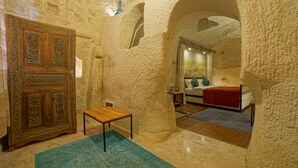 A room with a stone wall, a wooden cabinet, a small table, and a bed with a red blanket. - Taru Cave Suites (Ürgüp)