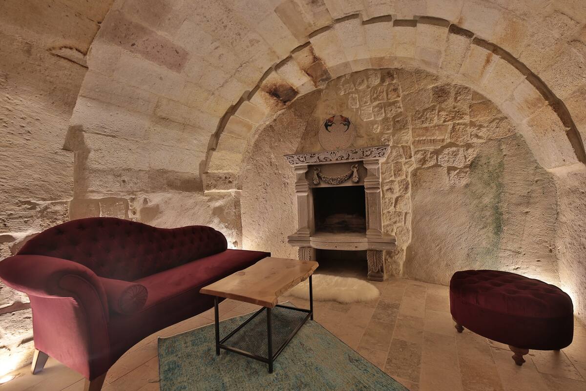 Une pièce aux murs de pierre, dotée d’une cheminée, d’une table en bois, d’un canapé rouge et d’un pouf rouge.