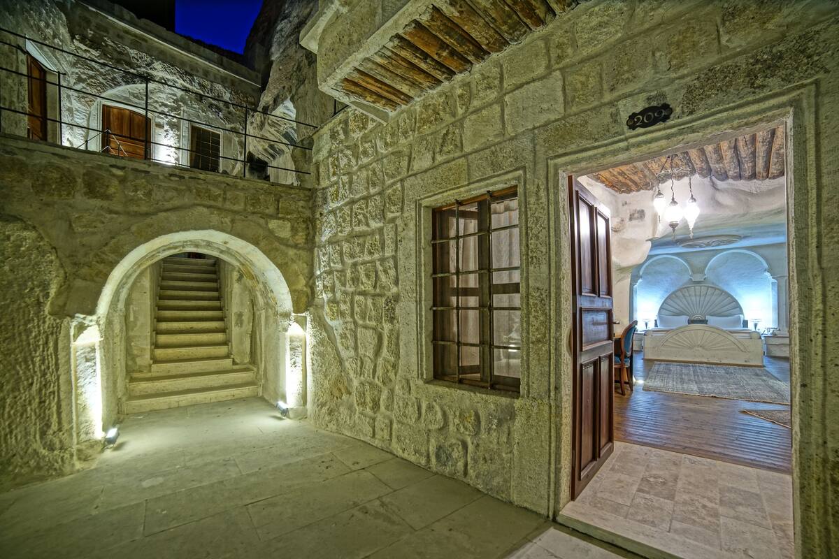Un couloir aux murs de pierre, avec un escalier menant à un balcon, un lit à baldaquin et une pancarte indiquant le numéro de la chambre.