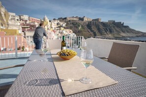 Una mesa con una botella de vino, un cuenco de fruta y una copa de vino en un balcón con vistas a una ciudad costera con edificios históricos.