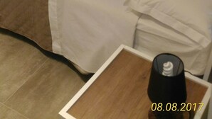 Una habitació amb paret de pedra, un llit amb roba de llit blanca, una petita taula de nit de fusta i una làmpada.