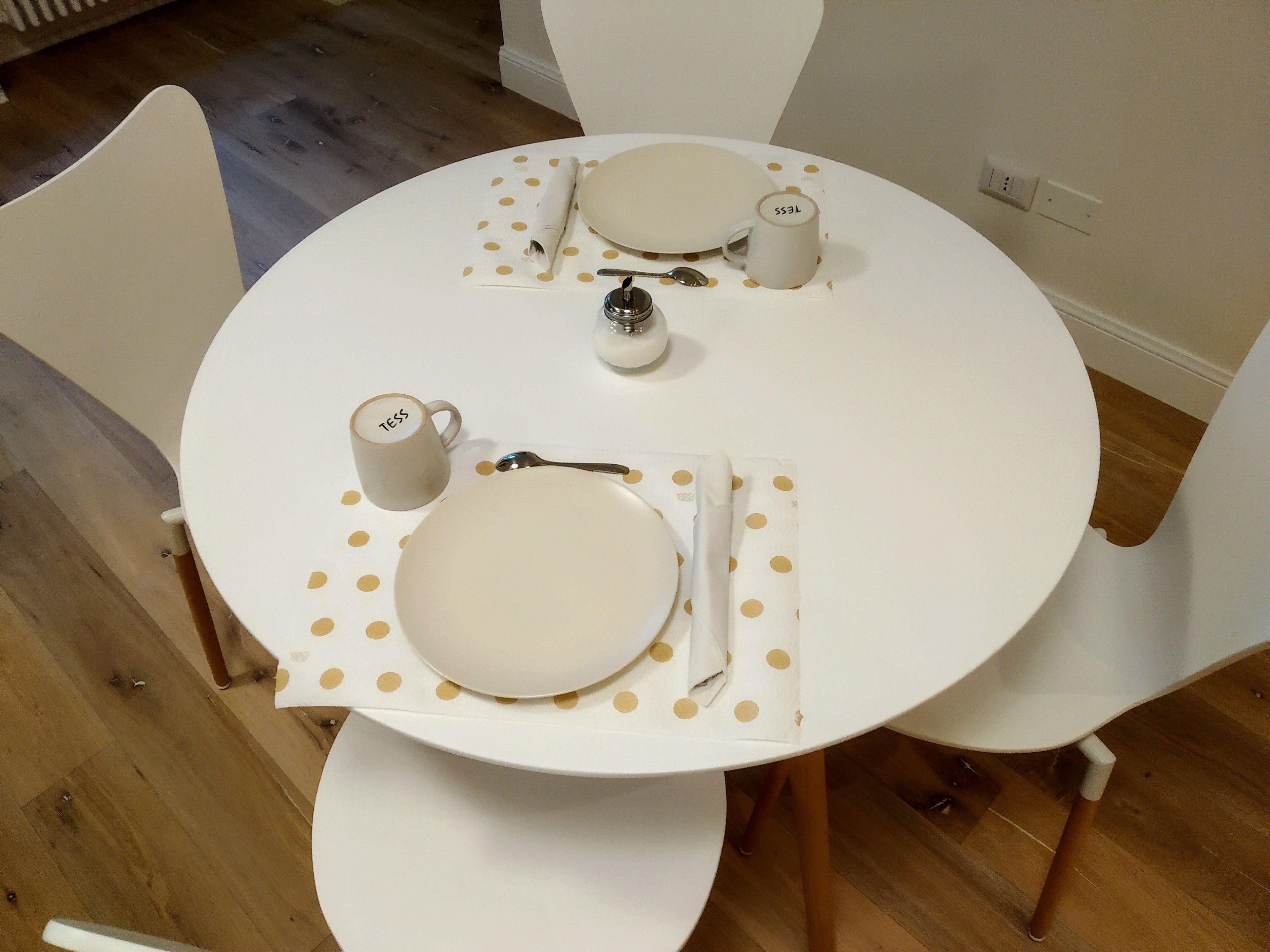A round dining table with white chairs, a white placemat with gold polka dots, a plate, a spoon, a cup, and a small container.