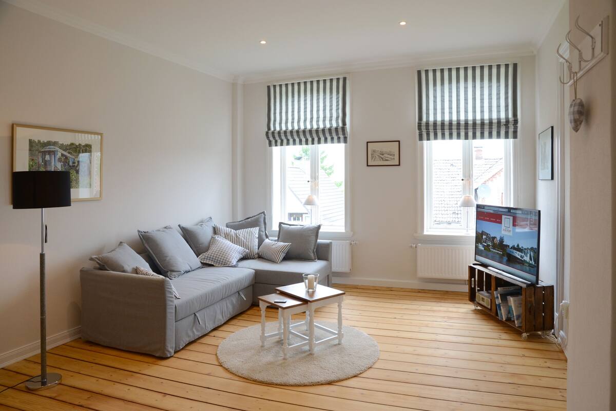A living room with a grey sofa, a small white table, a floor lamp, and a TV stand with books.