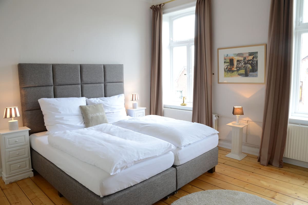 A double bed with a grey upholstered headboard, white bedding, two bedside tables with lamps, a window with curtains, and a framed picture on the wall.