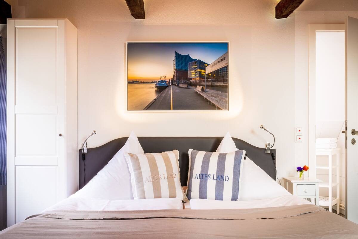 A neatly made bed with two pillows, a framed cityscape picture above it, and a small bedside table with a vase of flowers.