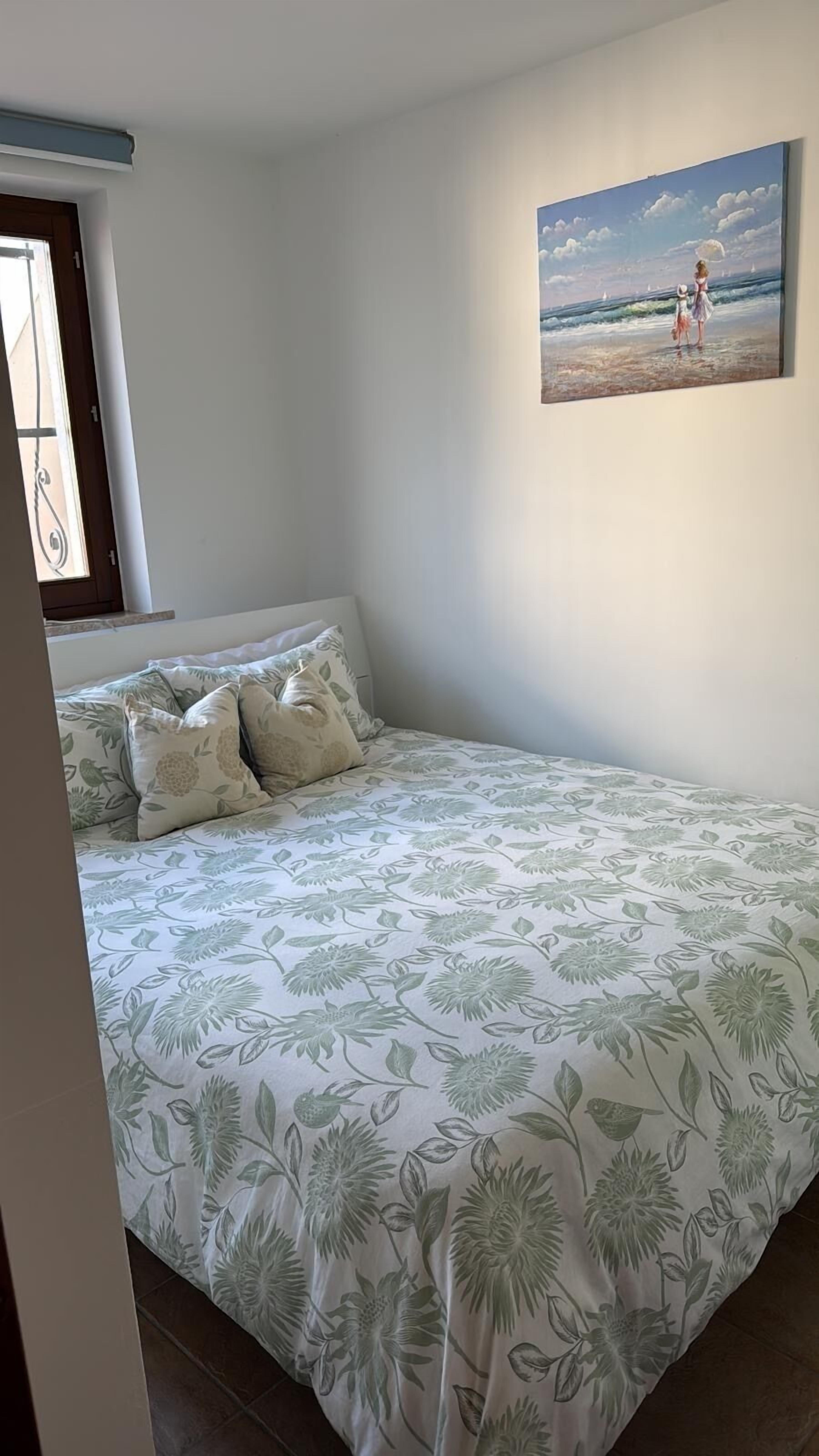 A bedroom with a bed, pillows, a painting of a beach scene, and a window with curtains.