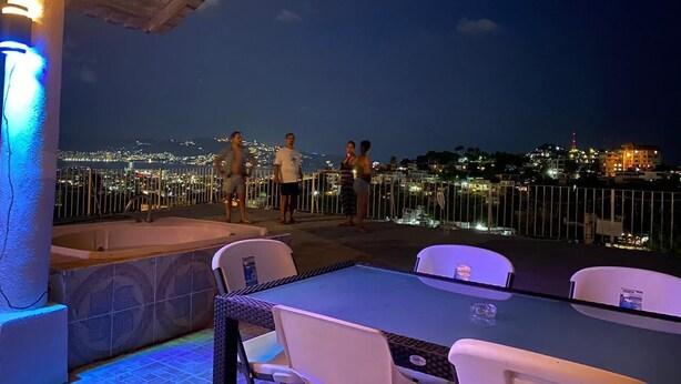 Une terrasse extérieure avec vue sur la ville, éclairée la nuit, comprenant une table et des chaises, une piscine et une rambarde.