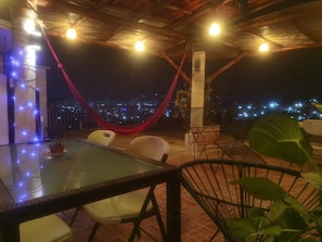 Uma área de estar externa com mesa de vidro, cadeiras, uma rede e vista para o horizonte noturno.