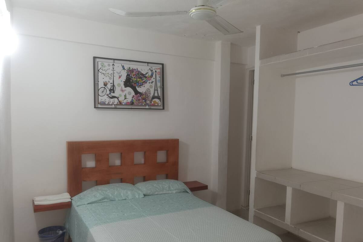 A bedroom with a bed, a nightstand, a wall-mounted picture, a ceiling fan, and a shelving unit.