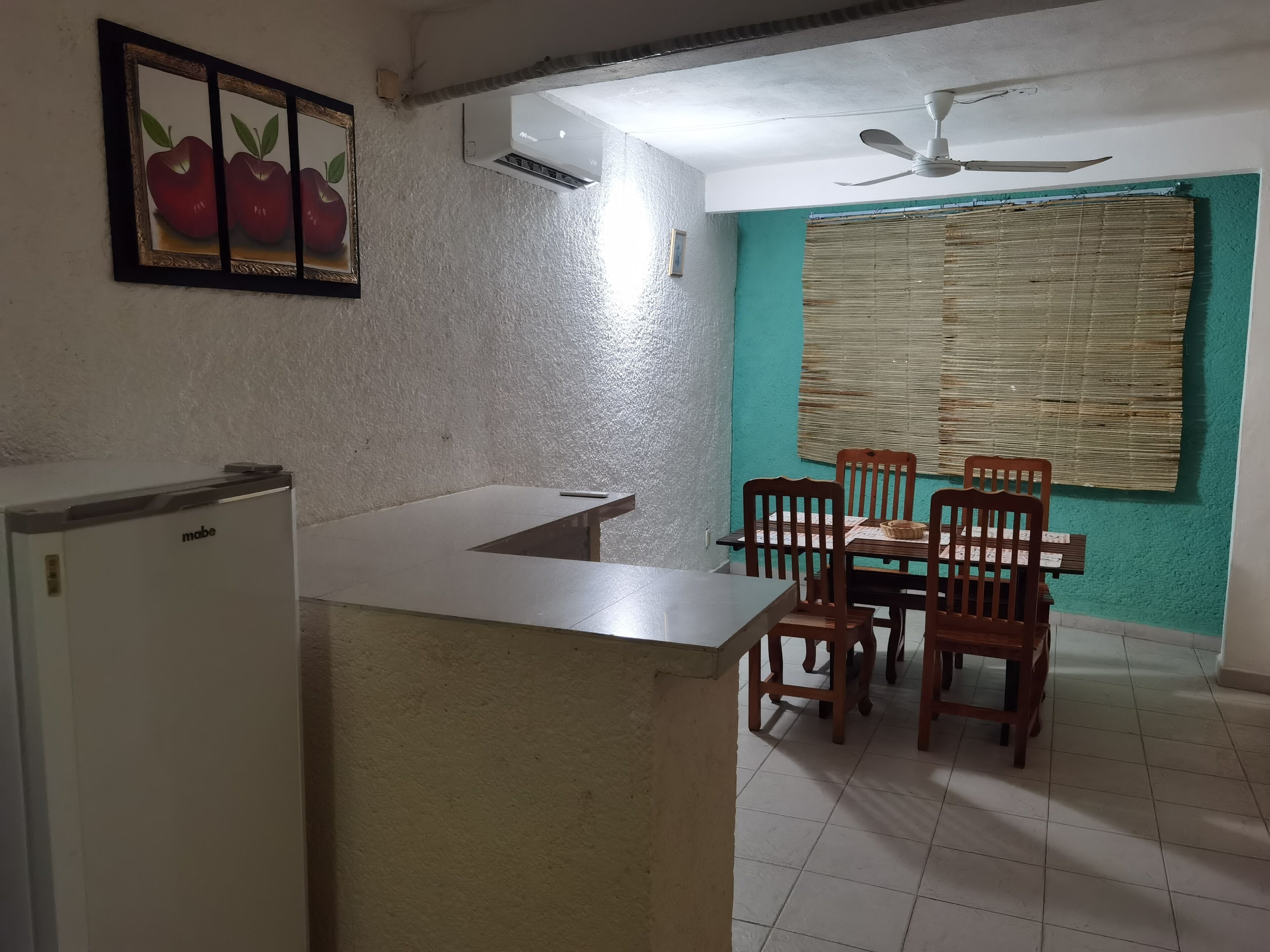 A kitchen with a white refrigerator, a wooden dining table with four chairs, a ceiling fan, and a wall-mounted air conditioner.