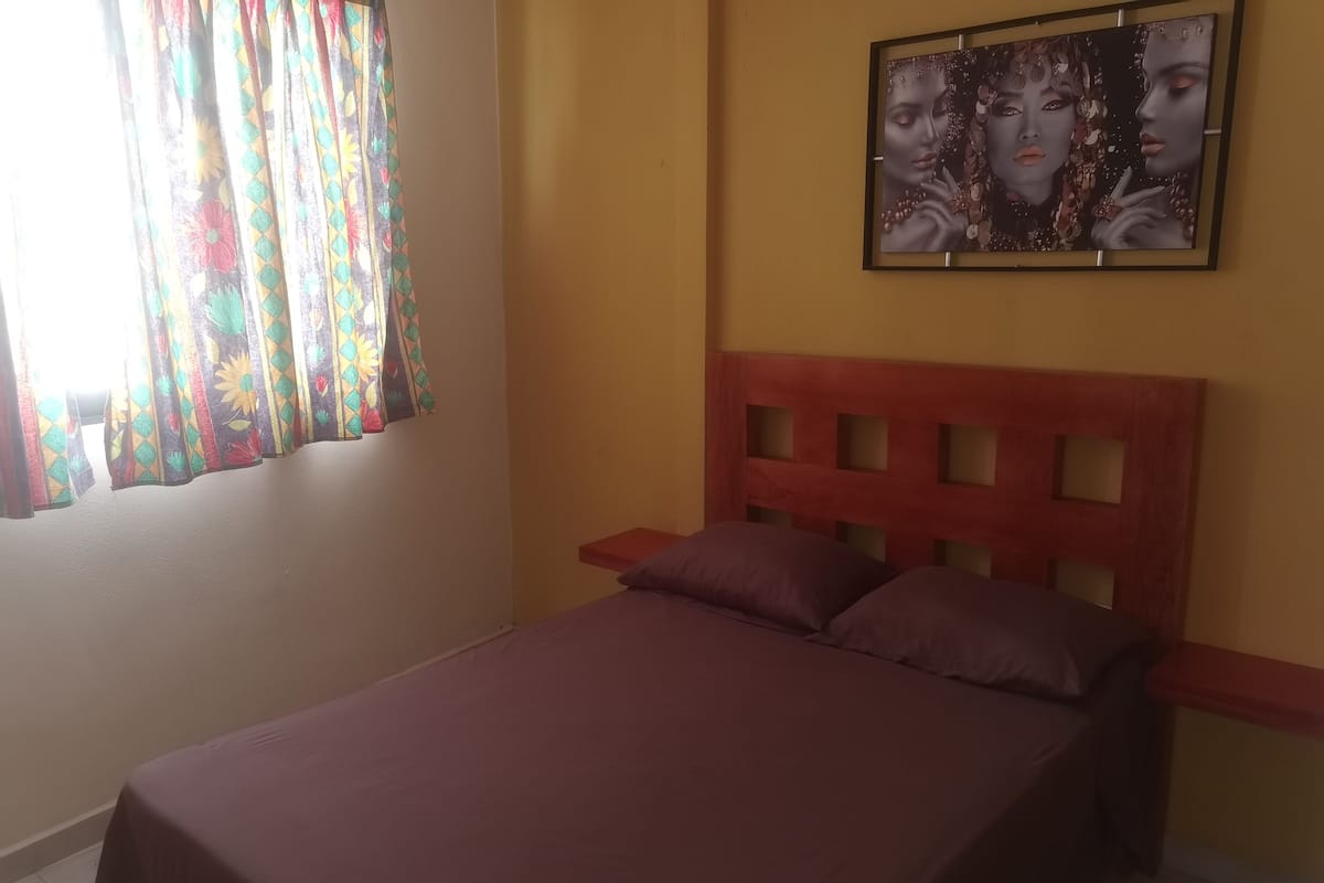 A bedroom with a bed, a nightstand, a wall-mounted painting, and a window with curtains.