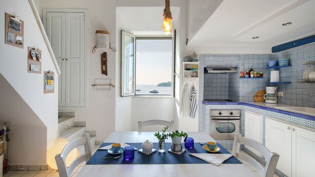 A dining area with a table set for two, a view of the sea through a window, and a kitchen with blue patterned tiles. - Corricella Sea Window (Procida)