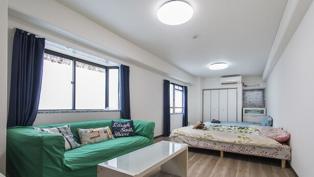 A compact room with a green sofa, a bed with floral bedding, a small table, and a window with curtains. - Shinsaibashi Su Casa Apartment (Osaka)