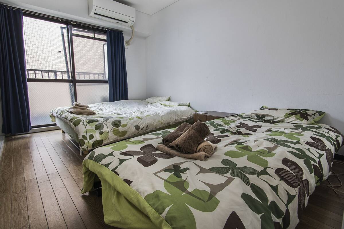 Ein Hotelzimmer mit zwei Betten, einem Fenster mit Vorhängen, einer Klimaanlage und Holzboden.