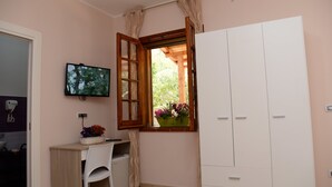Una stanza con una scrivania, una sedia, TV, armadio, finestra con tende e una pianta in vaso.