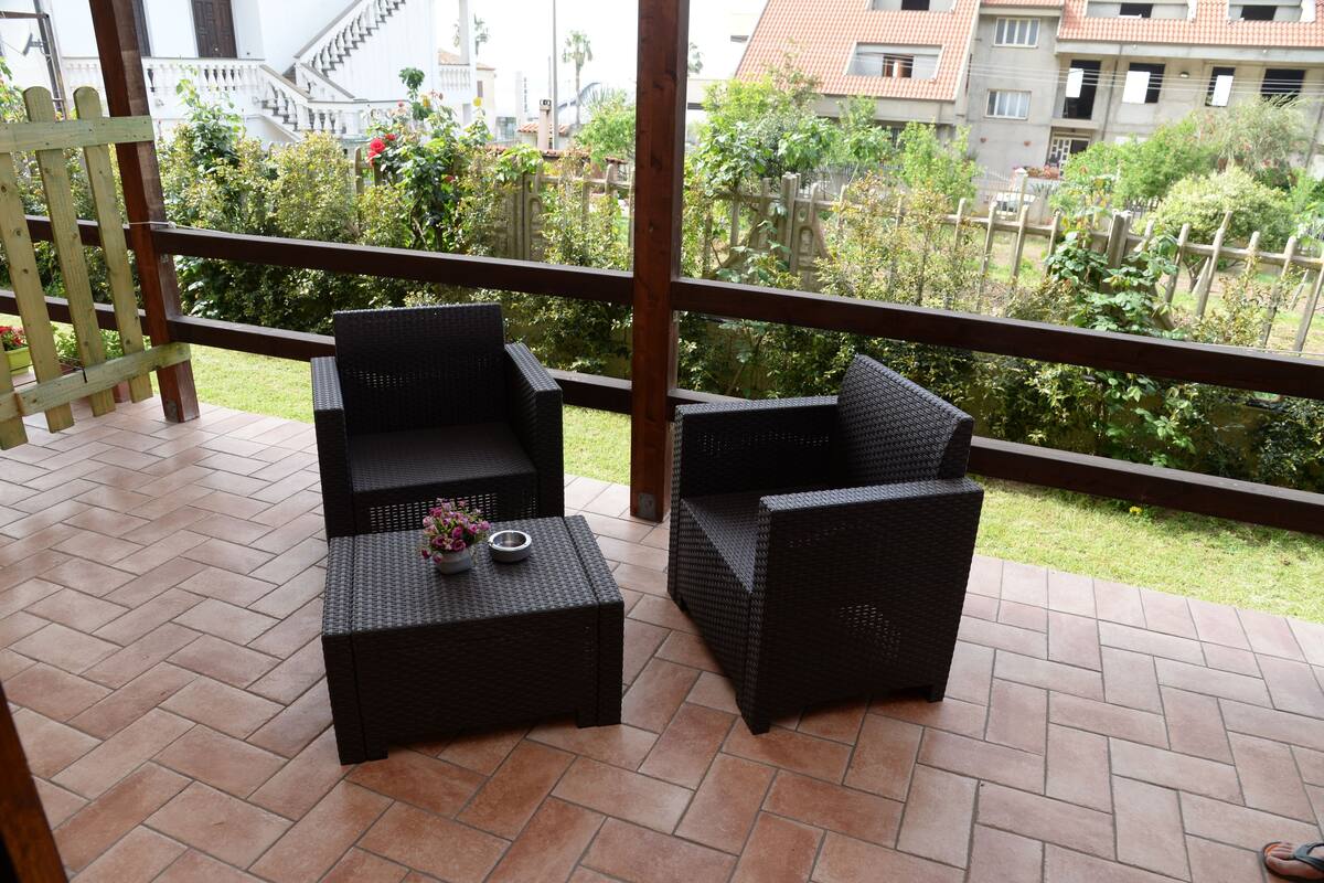 Ein Balkon mit Korbmöbeln und Fliesenboden, mit Blick auf einen Garten und Gebäude.