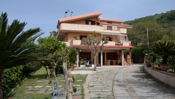 Una casa a due piani con un balcone, circondata da un giardino con palme e una statua.