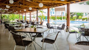 A covered outdoor dining area with tables and chairs, a pool in the background, and a view of buildings and greenery. - Hotel Peñon Suites (Girardot)