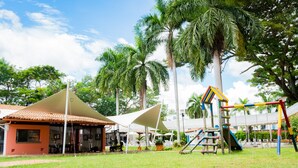 A playground with swings and slides, surrounded by palm trees and a building with a tiled roof. - Hotel Peñon Suites (Girardot)