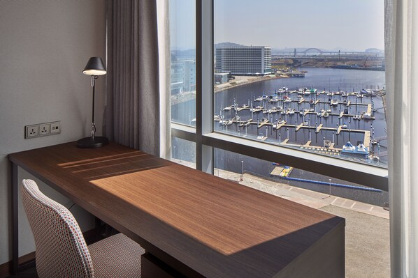 Ein Hotelzimmer mit Schreibtisch aus Holz, einer Lampe und Blick auf einen Hafen mit Booten und einer Brücke.