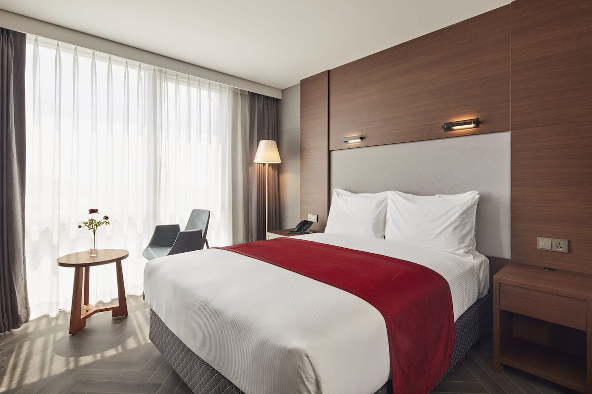 A hotel room with a large bed, a bedside table, a lamp, a chair, and a window with curtains.
