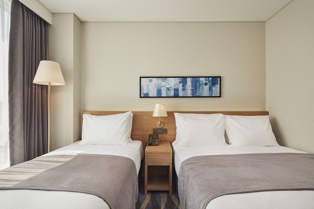A hotel room with two beds, a nightstand with a lamp, a painting on the wall, and a window with curtains.
