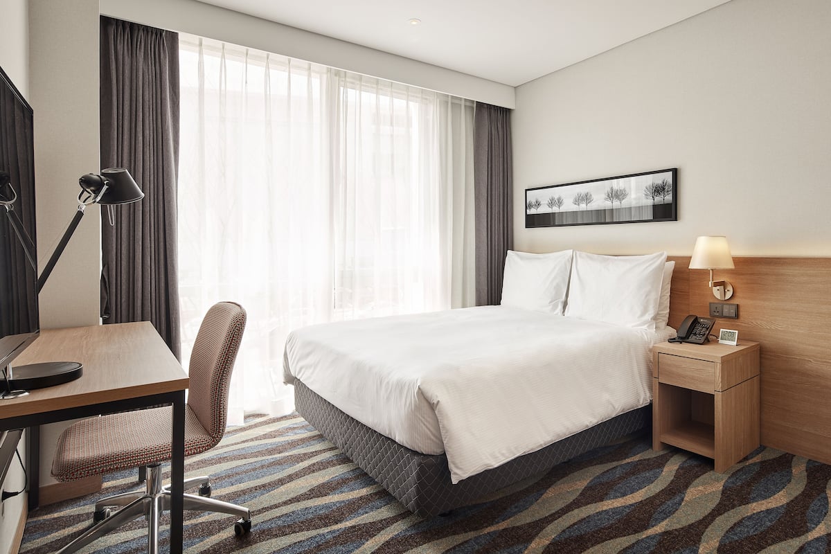 A hotel room with a bed, a desk, a chair, a nightstand with a lamp, and a window with curtains.