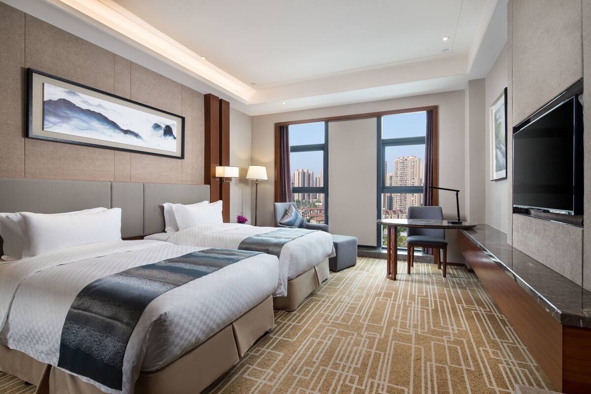 A hotel room with two beds, a desk, a TV, and a large window with a city view.