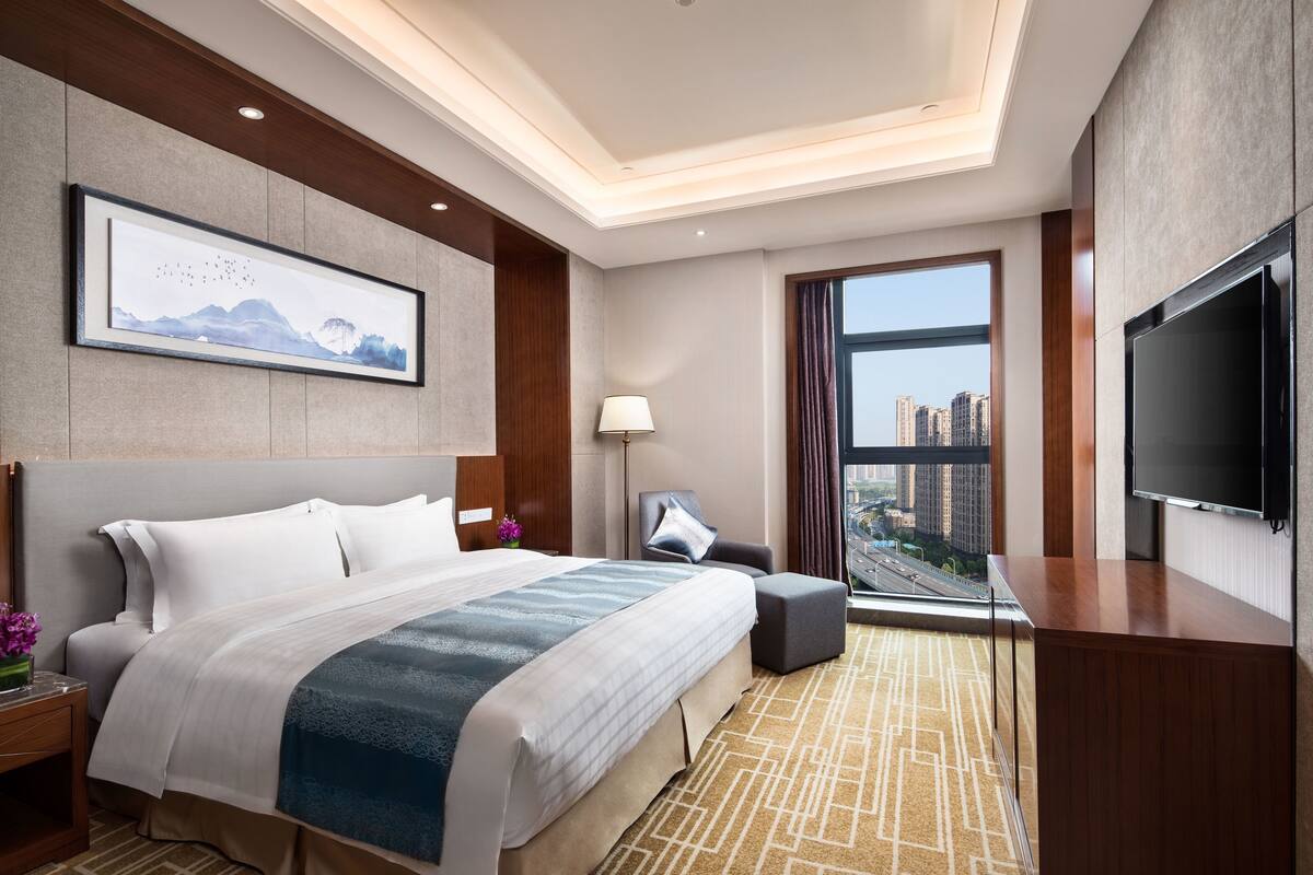 A modern hotel room with a large bed, a flat-screen TV, a desk, and a view of the city.