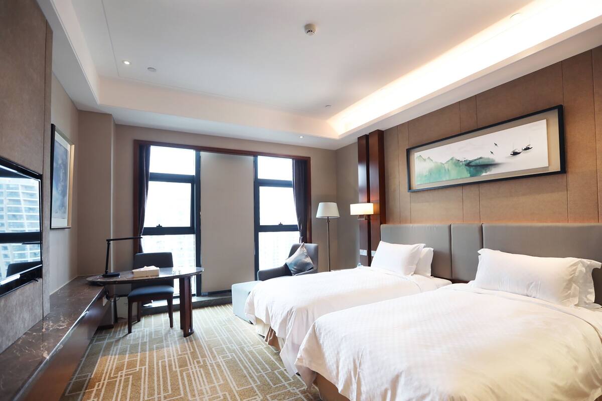 A hotel room with two beds, a desk, a chair, a TV, and a large window.