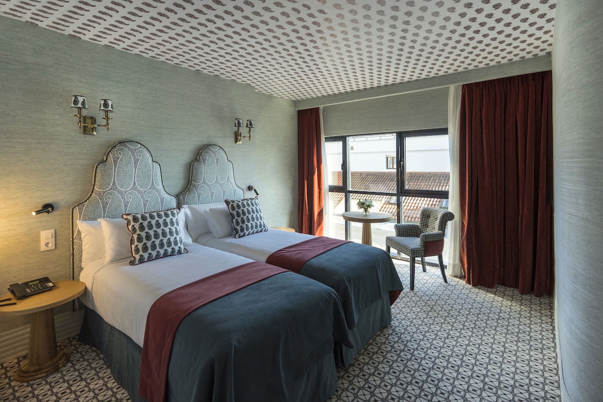 A hotel room with two beds, a small table, a chair, and a balcony with curtains.