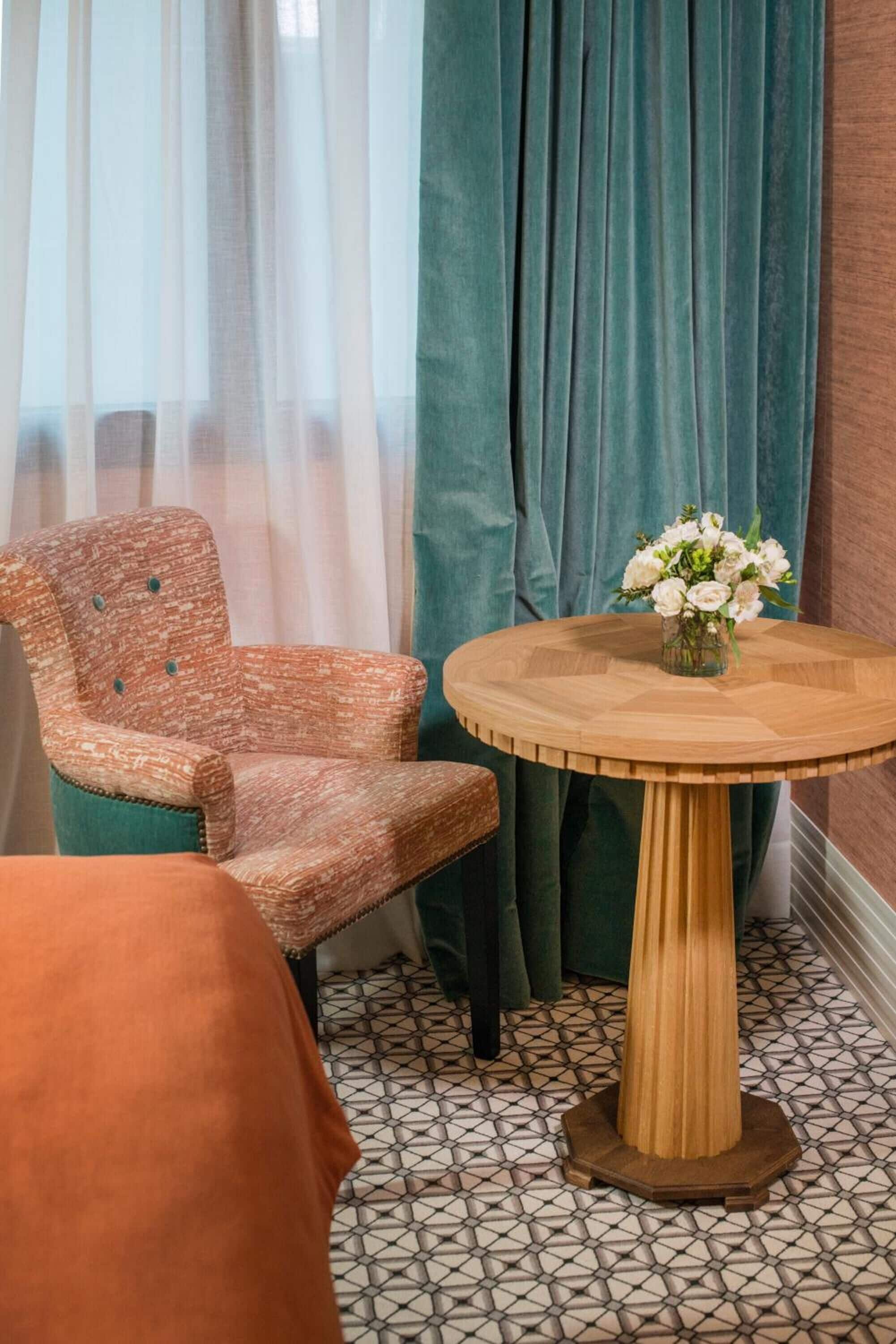 A room with a patterned armchair, a round wooden table with a vase of flowers, and a patterned floor.