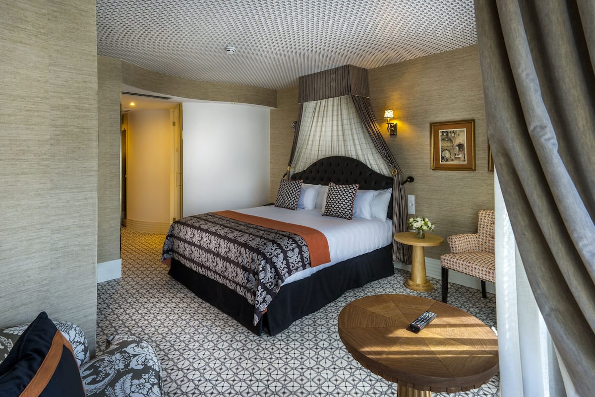 A hotel room with a large bed, a round wooden side table, a chair, and a small round table with a remote control.