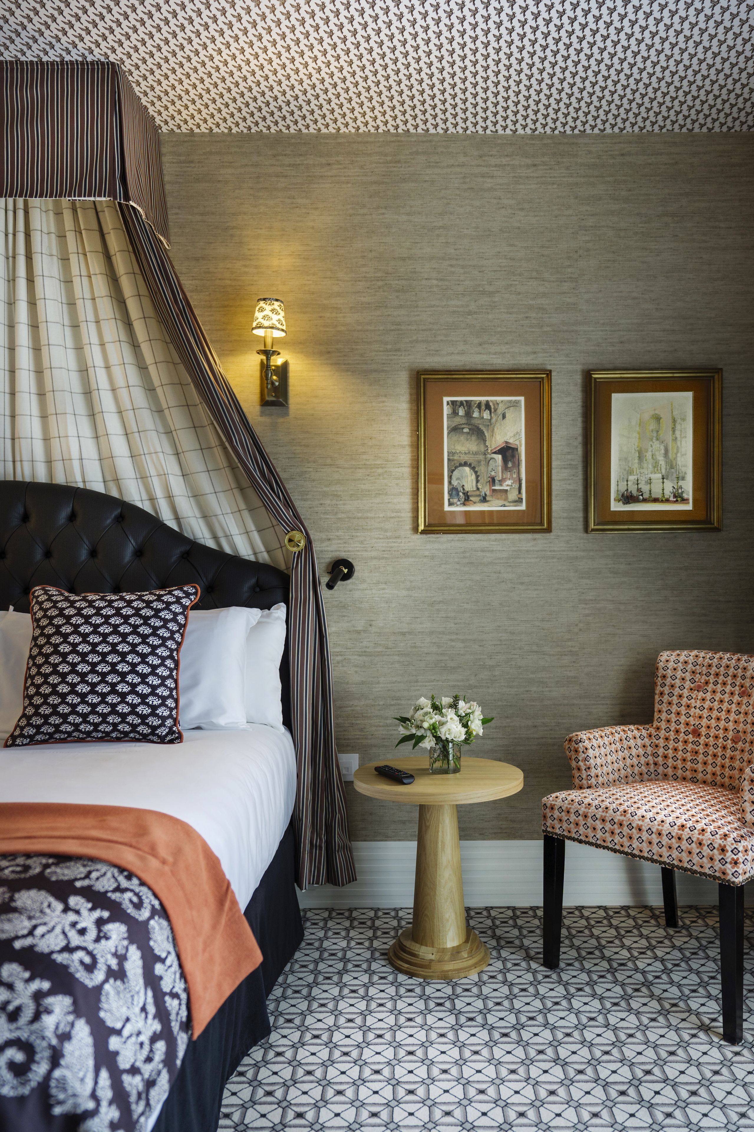 A hotel room with a tufted headboard bed, a round wooden side table, a patterned armchair, and two framed artworks on the wall.