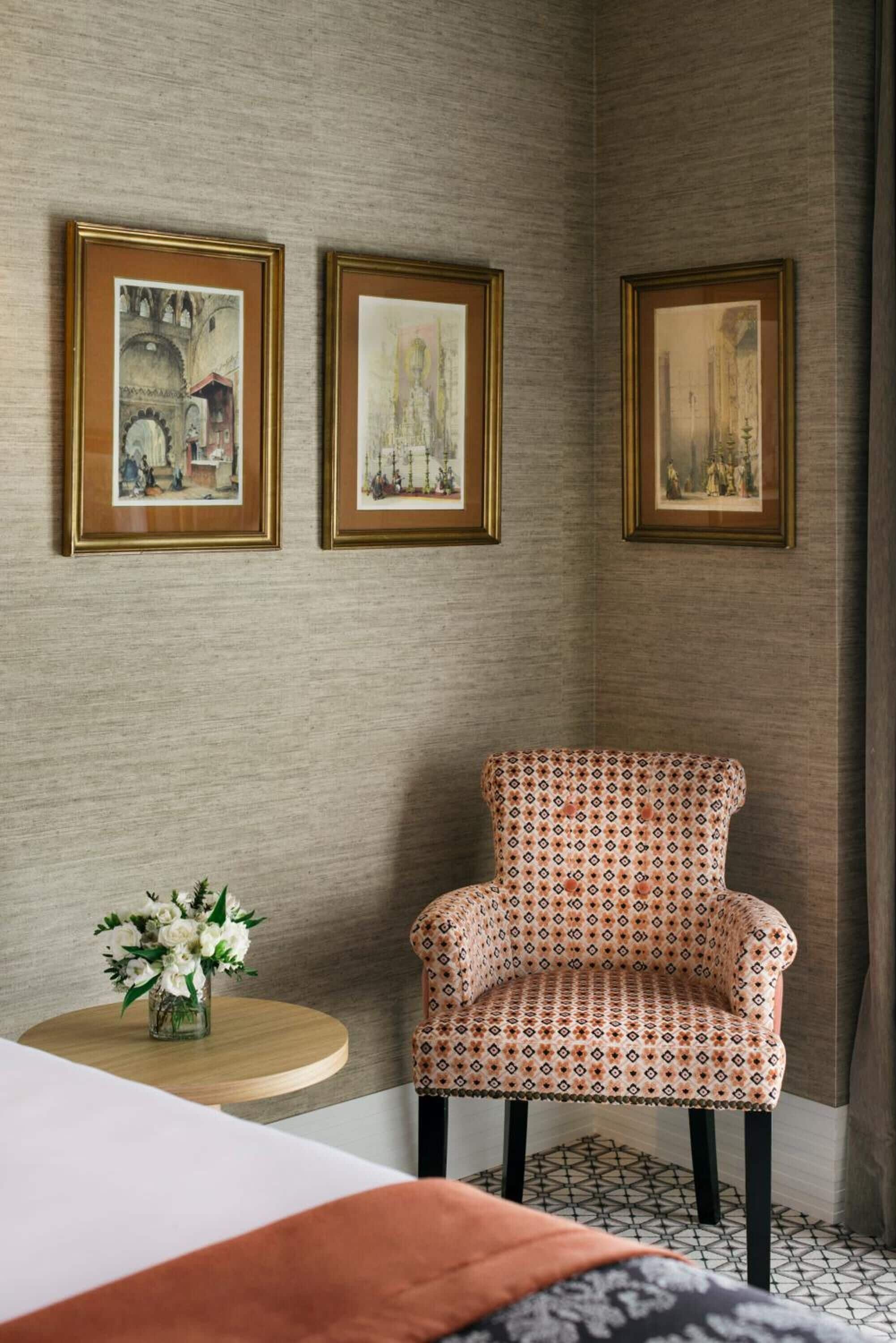 Una habitación de esquina con un sillón estampado, una mesita redonda con un jarrón de flores y tres cuadros en la pared.