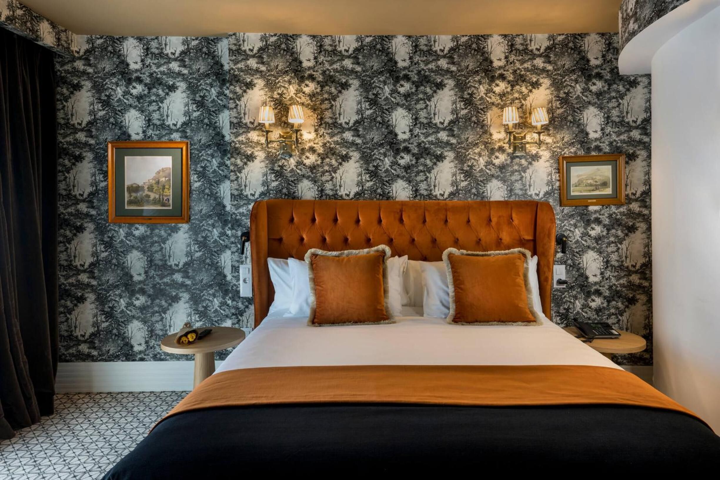 A bedroom with a tufted headboard, a bed with white linens and brown pillows, a round bedside table, and a wall with a forest-themed wallpaper.