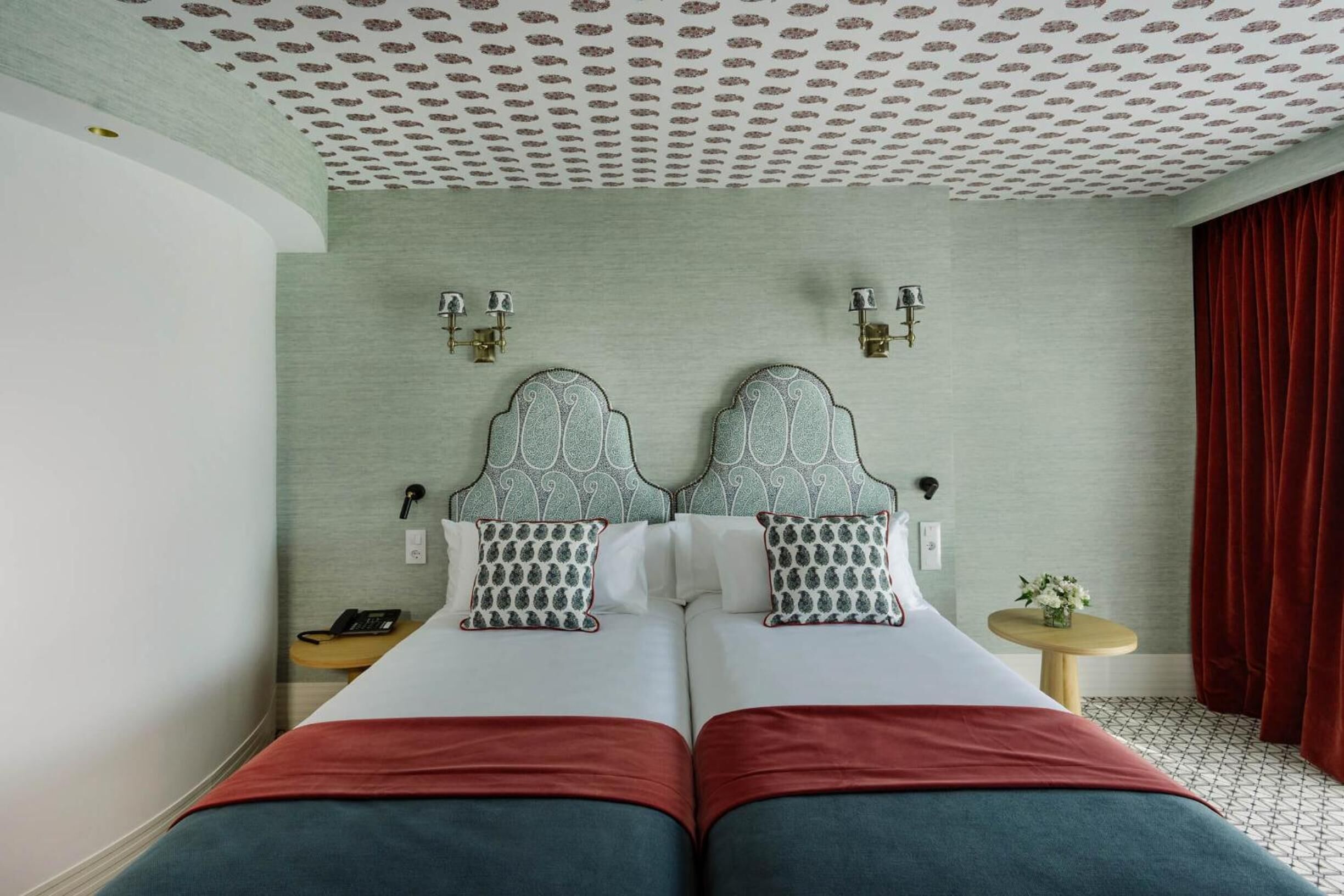 A double bed with a headboard, two bedside tables, a small round table with a vase, and a red curtain.