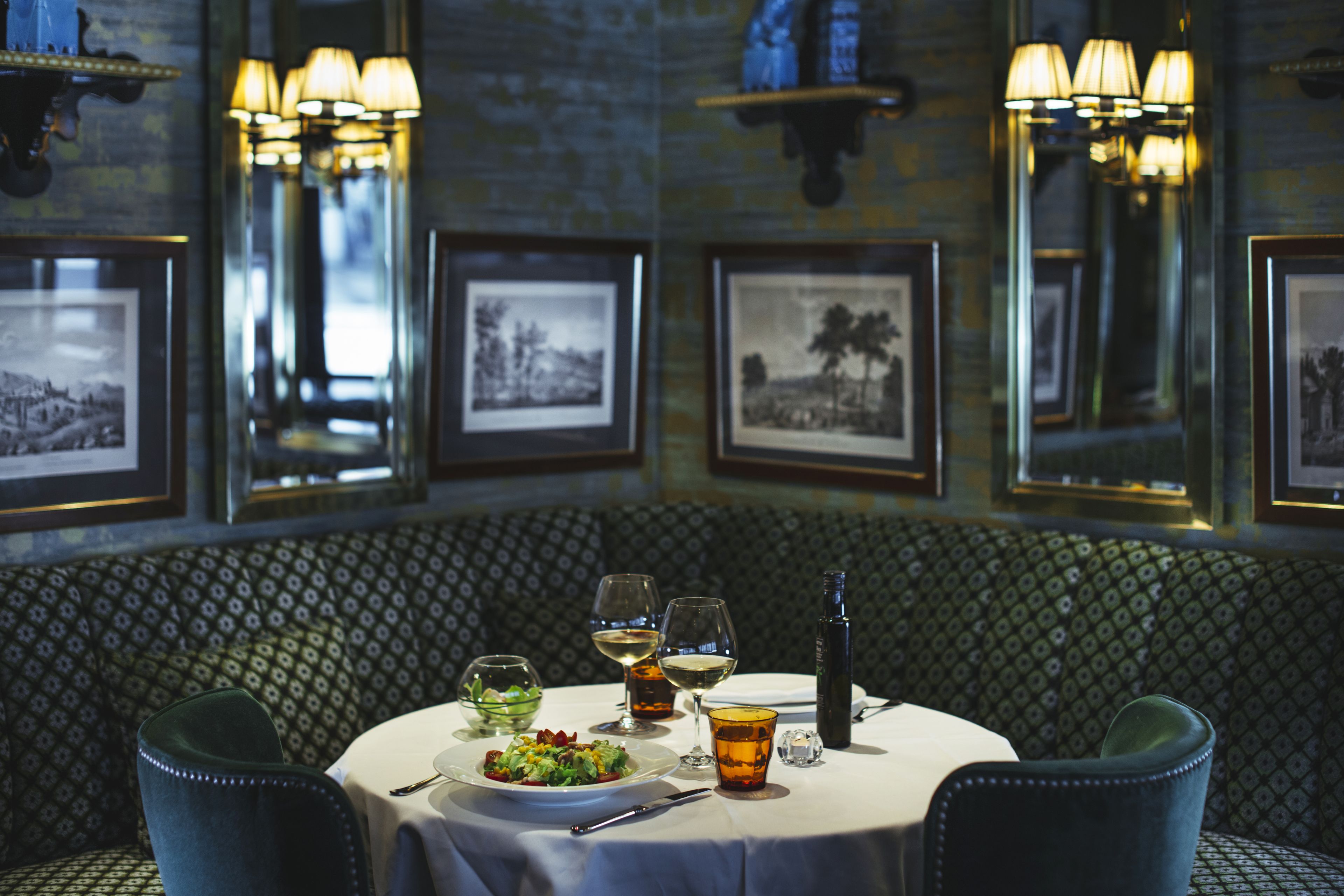 A dining area with a round table set for two, featuring a plate of food, glasses, and a bottle. The room has patterned wall upholstery and framed pictures on the walls.