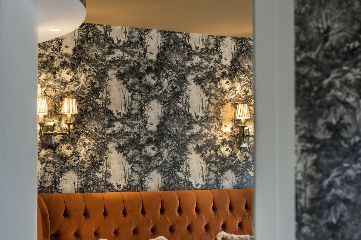 A bedroom with a tufted headboard, a patterned wallpaper, and two wall-mounted lamps.