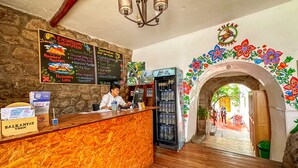 A hotel reception area with a wooden reception desk, a chalkboard with service information, a refrigerator stocked with beverages, and a colorful archway leading to the entrance. - Tucan Hostel (Cusco)