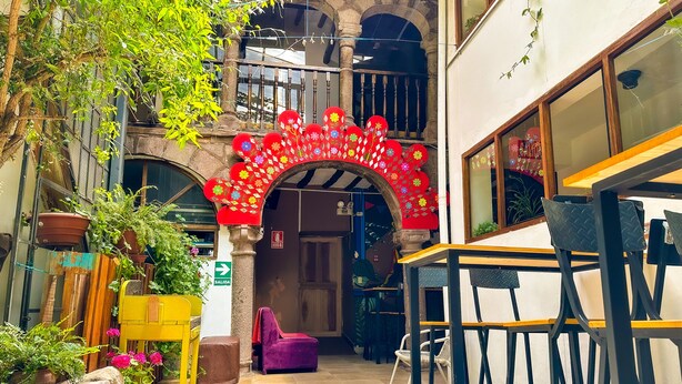 An outdoor seating area with tables and chairs, a stone archway with colorful decorations, and potted plants. - Tucan Hostel (Cusco)