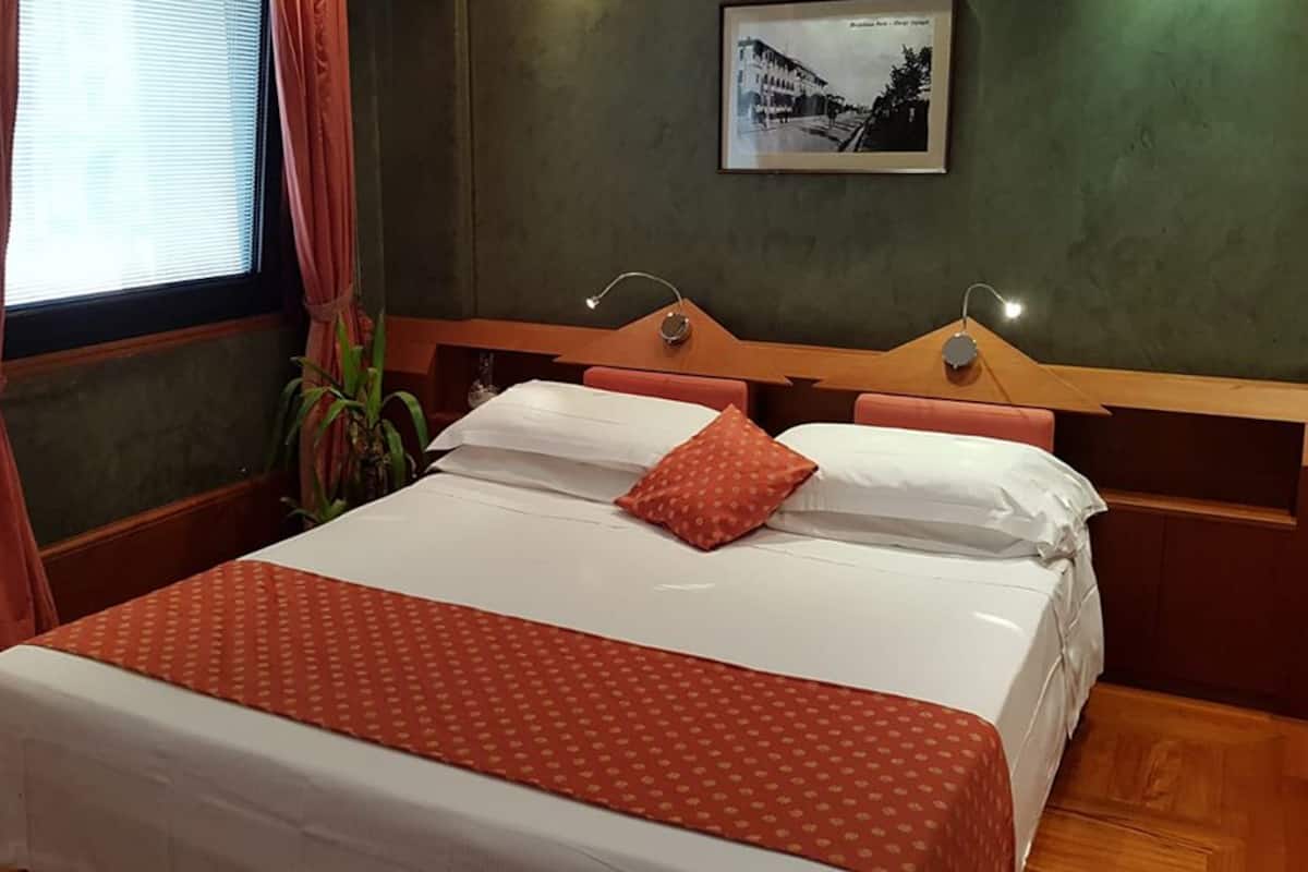 A hotel room with a bed, a wooden headboard, a nightstand, a wall-mounted lamp, and a framed picture.