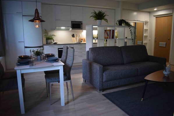 A modern kitchen with white cabinets, a dining table with chairs, and a grey sofa in the living area.