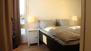 A bedroom with a bed, two bedside lamps, a nightstand, a vacuum cleaner, and a window with curtains. - DP Apartments Vaasa (Vaasa)
